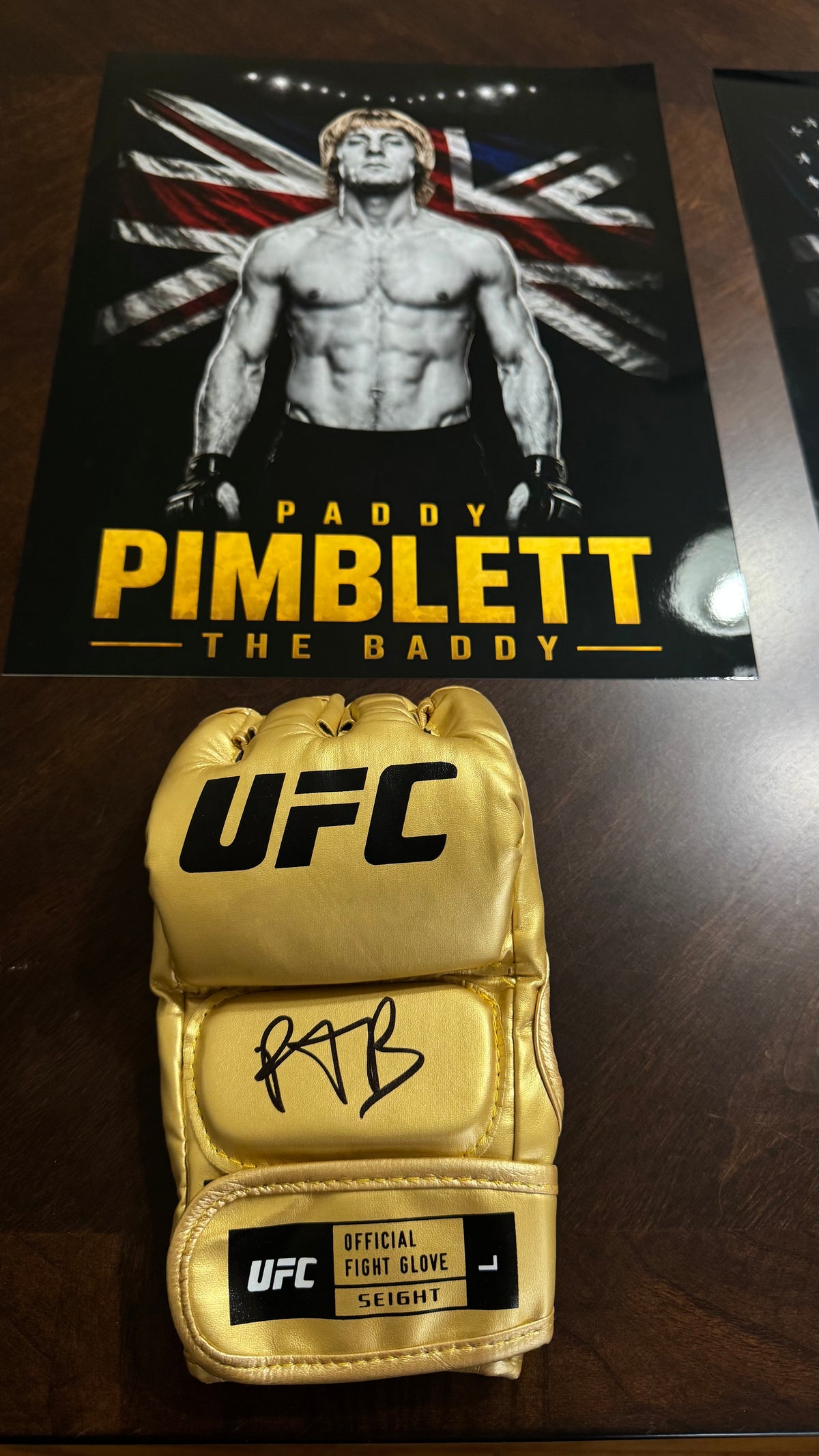 A gold UFC fight glove signed by Paddy Pimblett, displayed on a dark wooden surface in front of a Paddy Pimblett poster.