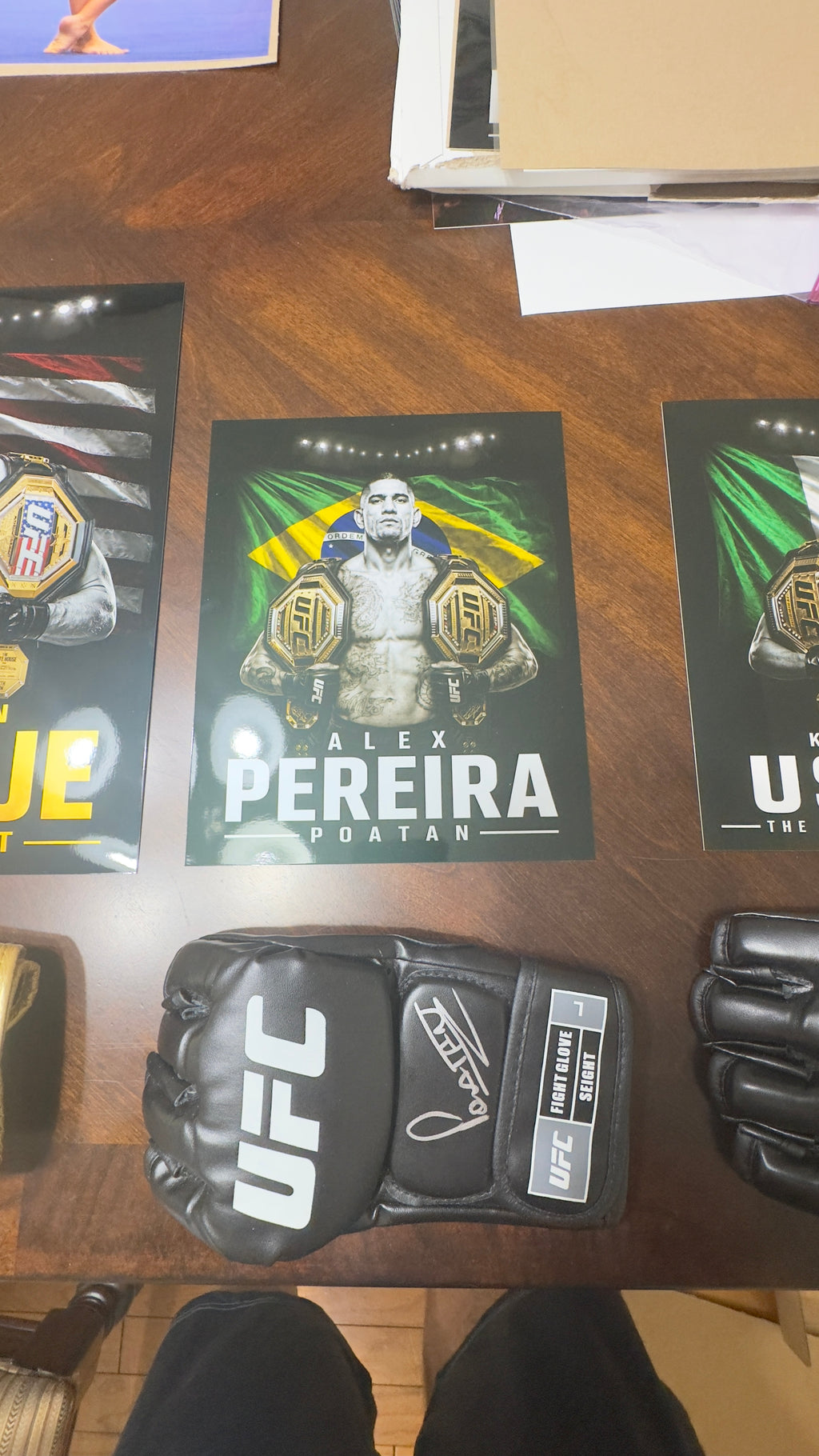 A signed black UFC glove is displayed on a wooden table, surrounded by Alex Pereira fight posters and another glove in the foreground.