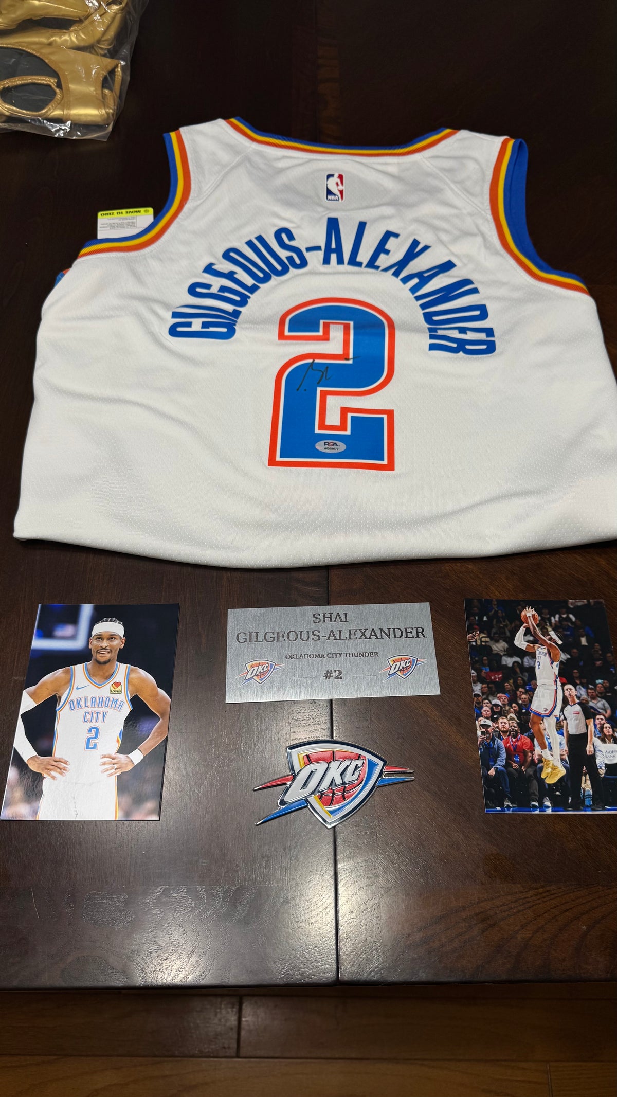 A white Oklahoma City Thunder basketball jersey with 'GIGGOUS-ALEXANDER' and number 2 on the back, displayed on a dark wooden table with two player photos, a signed plaque, and an OKC logo.