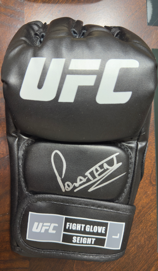 A black UFC fight glove with a white printed UFC logo, a silver autograph on the palm, and a white-and-gray size label reading 'FIGHT GLOVE SEIGENT' on a wooden surface.